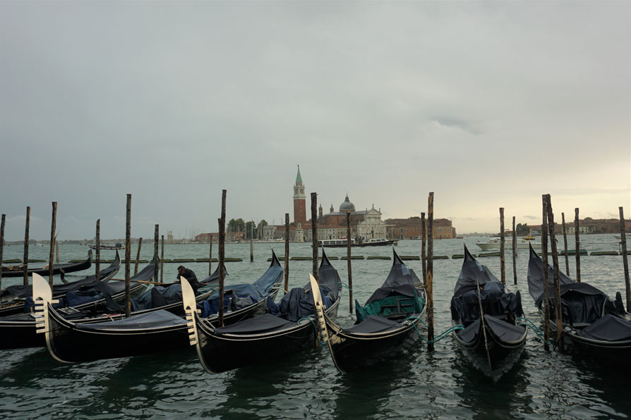 Peaceful photo of a row of empty boats, grey water and sky, European buildings in the distant background.