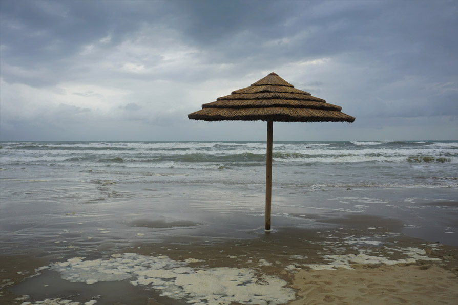A grey, desolate sky over frothy, stormy waves.
		In the foreground, an abandoned beach, a lone straw-thatched permanent beach umbrella.
		In front of the umbrella, mix of sand and pools of ice.