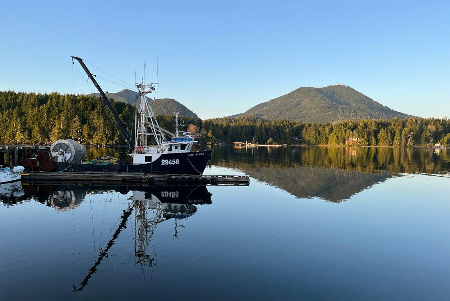 Photo is captioned, 'Ucluelet, Vancouver Island'. Image shows a black and white fishing boat with a crane type apparatus in the aft section.
		It is a side view of the boat, which is docked flush against a narrow plank. The reflection of the boat in the water is pristine, along with the forest, mountain, and clear blue sky behind the boat.