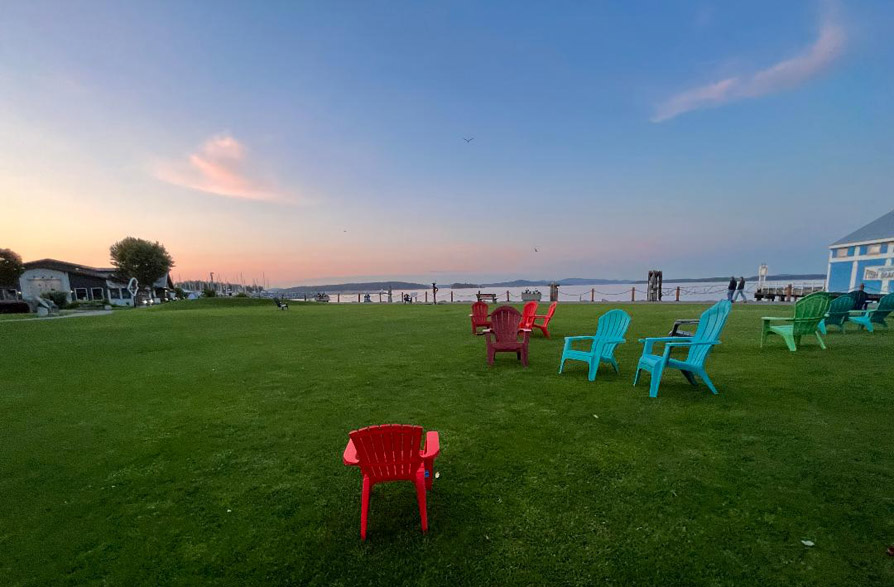 Photo is captioned, 'Sidney, Vancouver Island'. Image is of a sunset scene; red, blue, and green empty lawn chairs are scattered on a lush green lawn.
		At the horizon is a body of water, and beyond it a dark line of the land beyond the water. Above the water is rose and blue coloured sky,
		mostly clear except for a few wistful clouds.