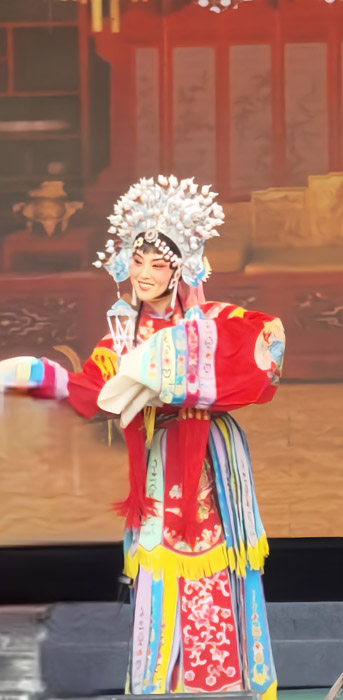 Photo is uncaptioned.
		Image shows a performer dressed in elaborate and vibrant cultural garments.
		The garments are a mixture of red, white, light blue, and yellow.
		The performer wears an elaborate headress with her arms raised as if dancing.