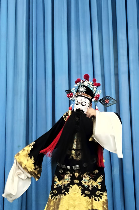 Photo is uncaptioned.
		Image shows a performer dressed in elaborate and vibrant cultural garments.
		The garments are mainly black, white, and gold.
		The performer dances in front of a tall blue curtain, creating an appealing colourful scene.
		The performer wears an elaborate headress.