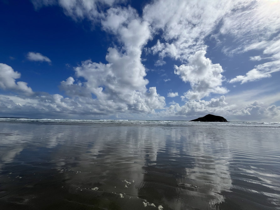 Photo shows a beach view of bright blue, cloud-filled reflected on wet brown sand.
			Close to the horizon the distant surf of waves can be seen. 
			On the right side of the screen, at the horizon, a small island, Incinerator Rock, is silhouetted, a shape like a diamond sideways, half buried.