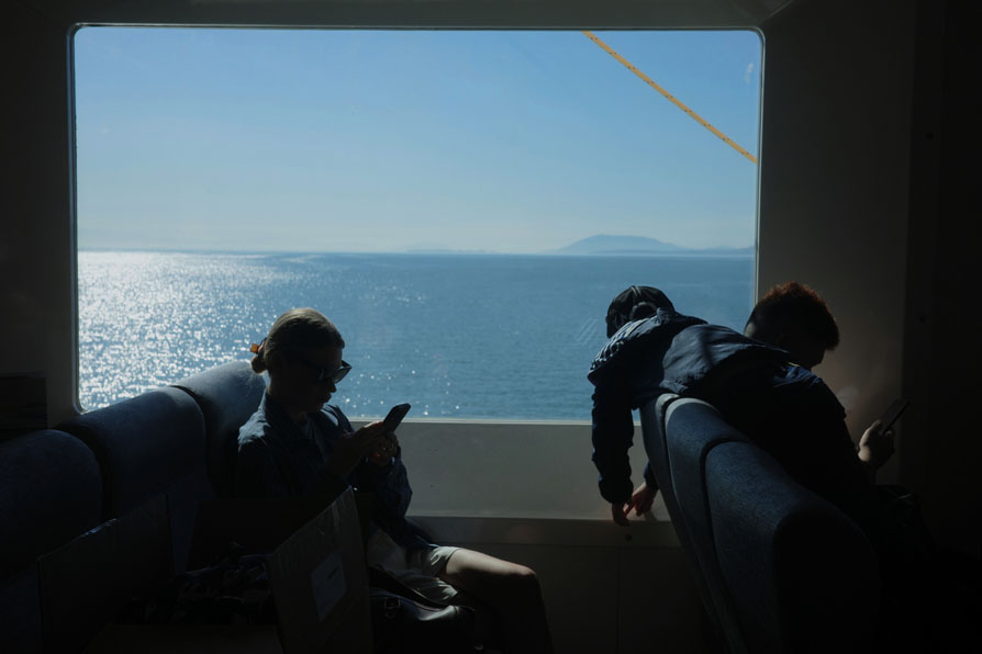 Photo shows the view out a ferry window. Silhouette of passengers,
	one a young person scrolling their phone. Another young person is laying on top of the 
	chairs, gazing out the window.